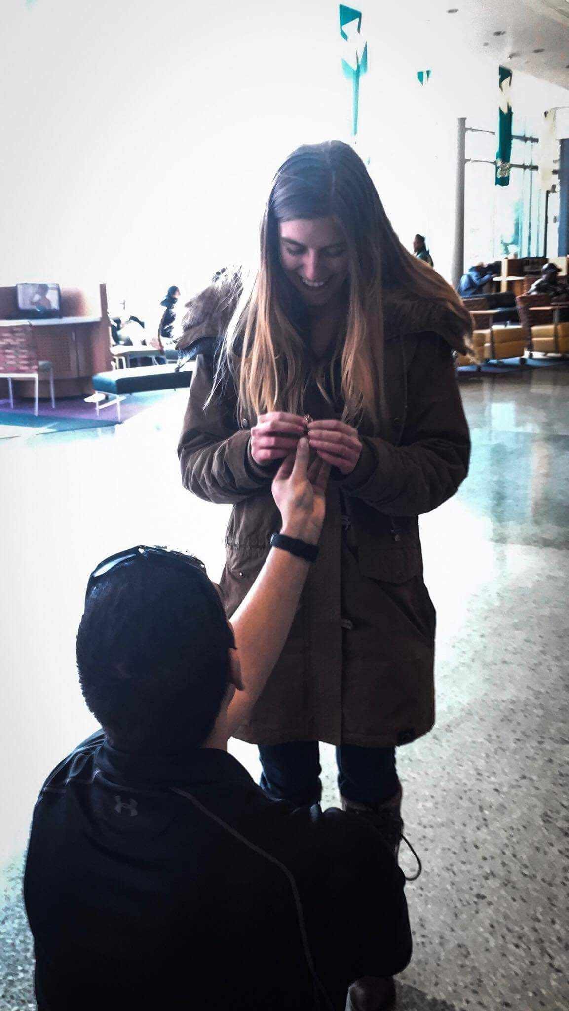 Person proposing to another person with a ring inside a Wayne State University building with large windows in the background.